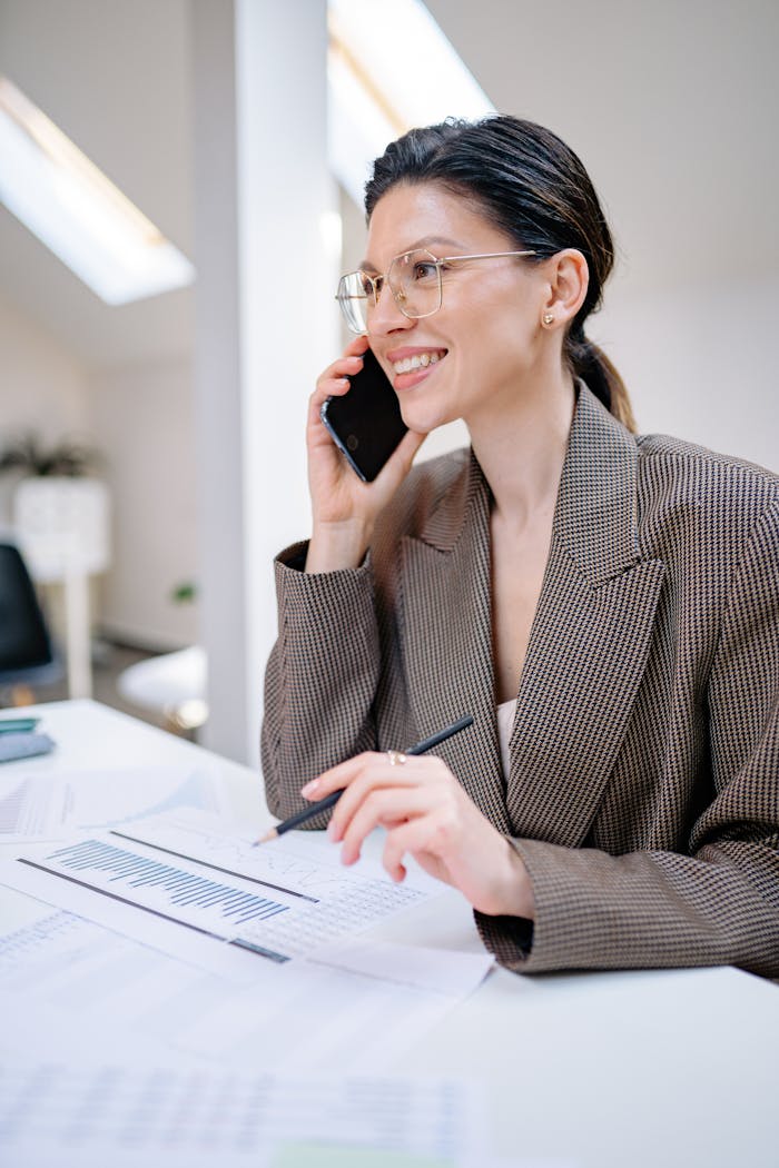 Confident businesswoman talking on smartphone while reviewing financial documents at her desk.