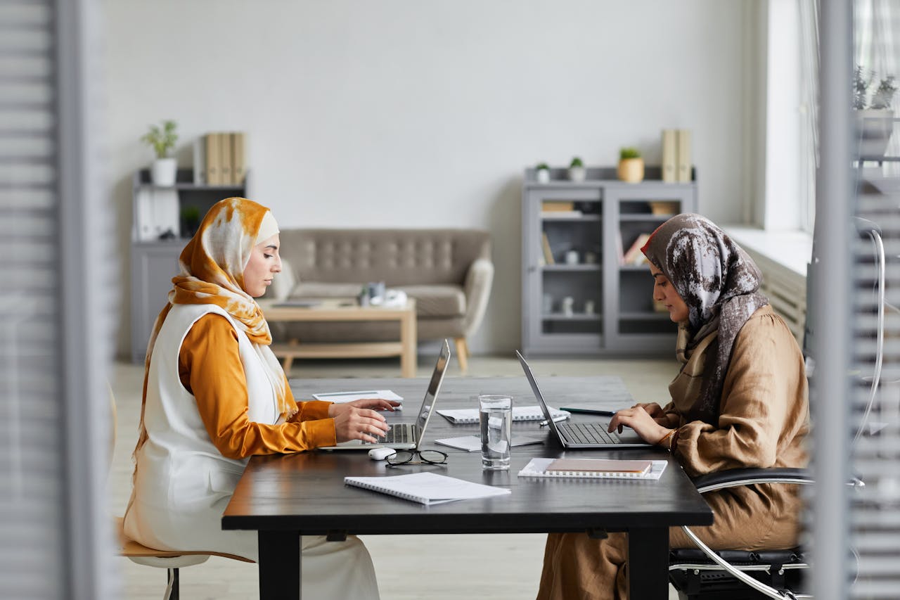 Two women wearing hijabs working on laptops at a modern desk in an office setting.